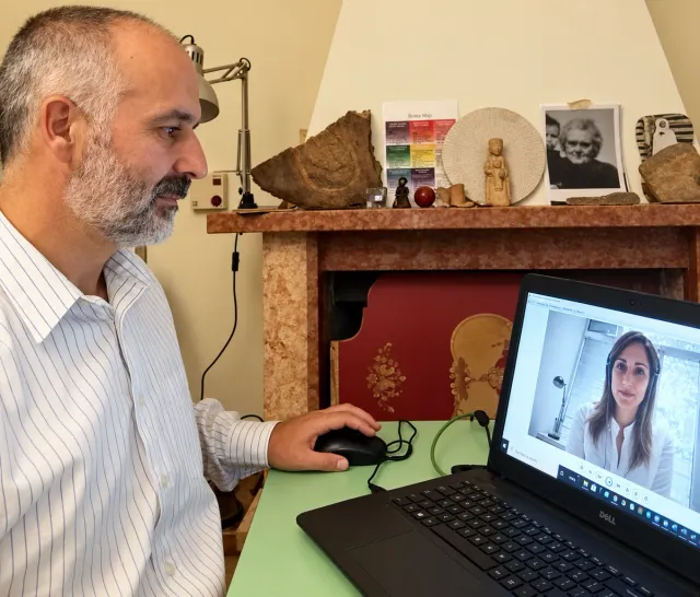 Iuri alla scrivania durante una videochiamata, con fotografie di famiglia e ricordi sul caminetto alle sue spalle
